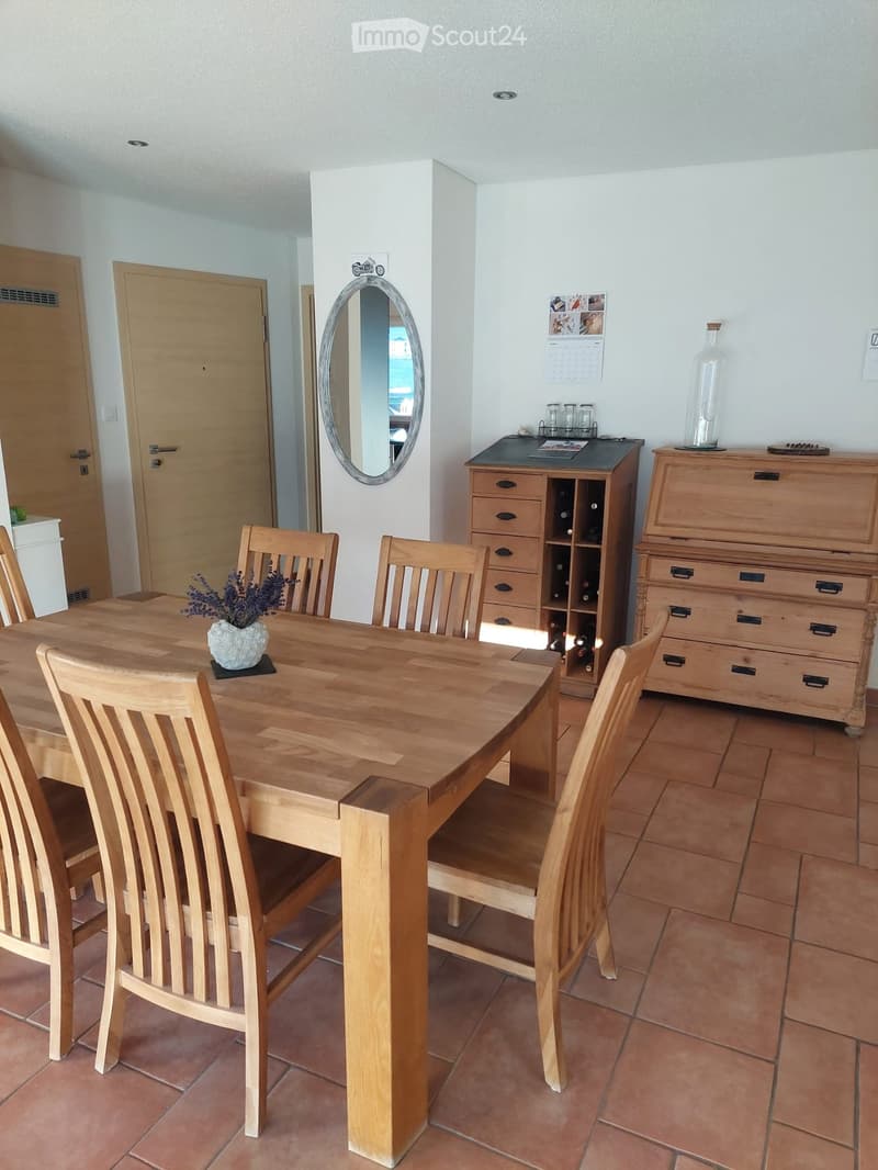 Dining room with a wooden dining table, six chairs, a wooden cabinet, a mirror, a vase with flowers, a calendar, and a bottle on a sideboard.