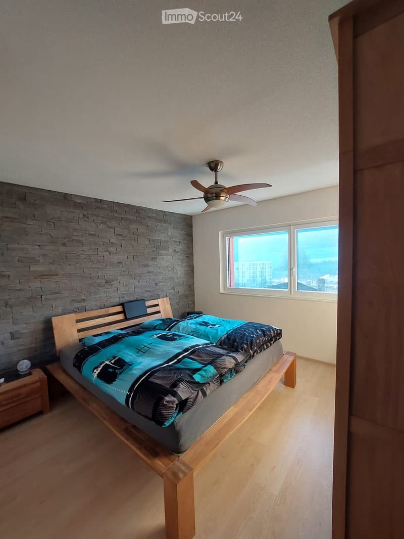 Modern bedroom with wooden bed, blue and black bedding, nightstand, and ceiling fan. Large windows offer a city view.