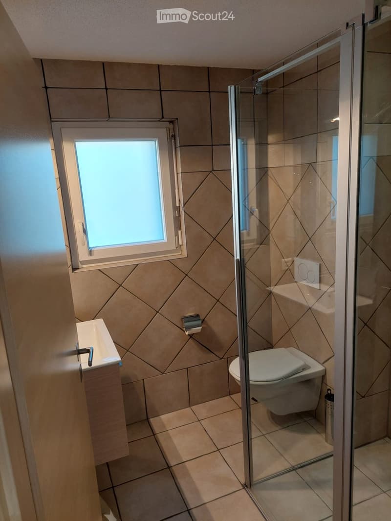 Modern bathroom with beige tiles, a glass-enclosed shower, a toilet, a sink with a modern faucet, and a window for natural light.