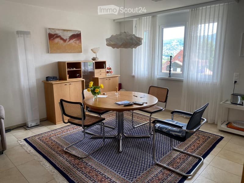 Dining area with a round wooden table, four chairs, a cabinet, a vase with yellow flowers, and a painting on the wall.