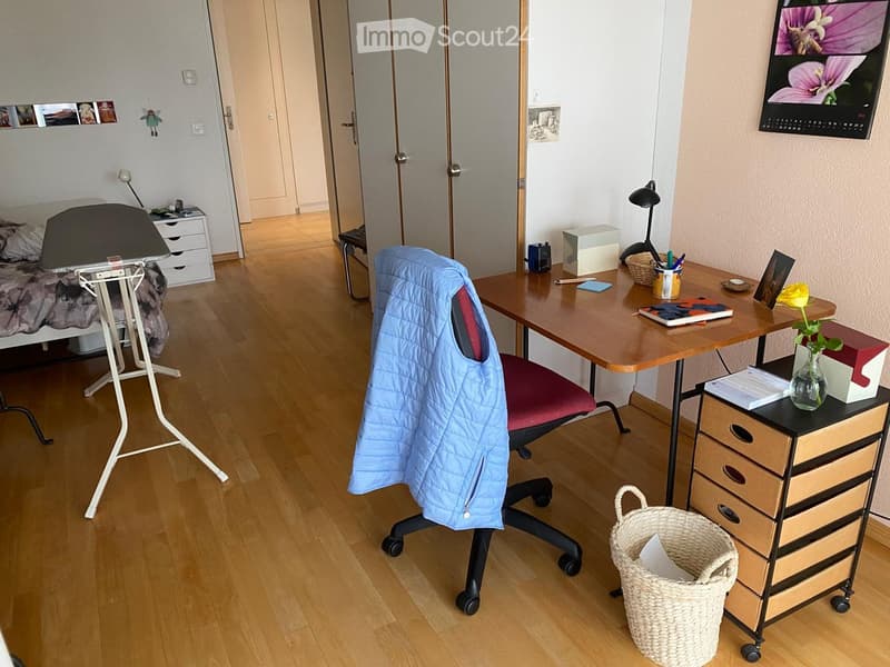 A well-lit room with a wooden floor, a desk with a chair, a drawer unit, a wicker basket, a flower vase, a screen, a lamp, a bed with a nightstand, a mirror, and a picture frame.