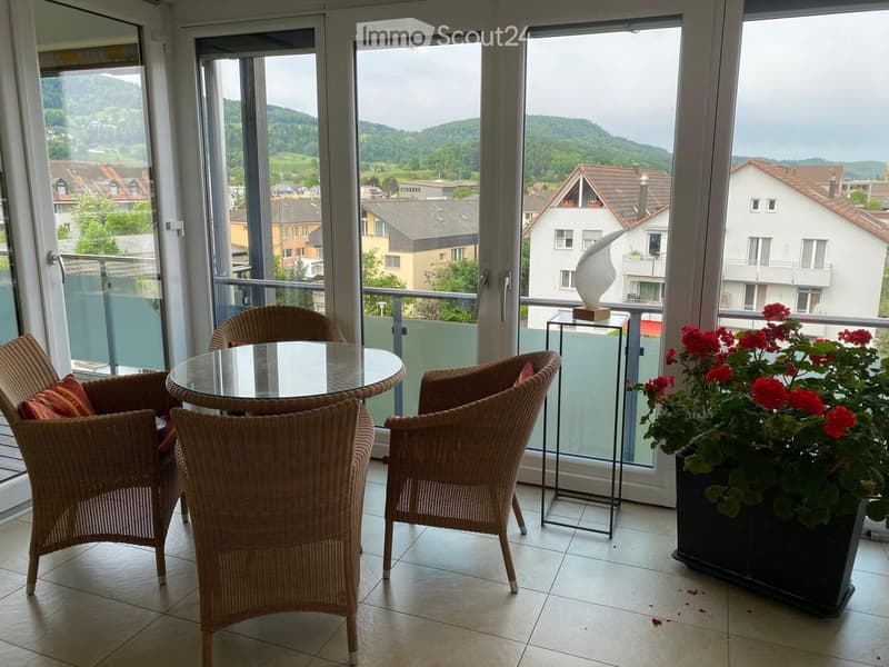 Balcony with a glass table, wicker chairs, and a flower pot with red flowers, overlooking a scenic town and hills.