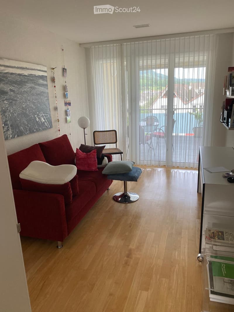 Living room with wooden floor, red sofa, white and red cushions, a small round table, a floor lamp, and a view of a balcony with outdoor furniture.