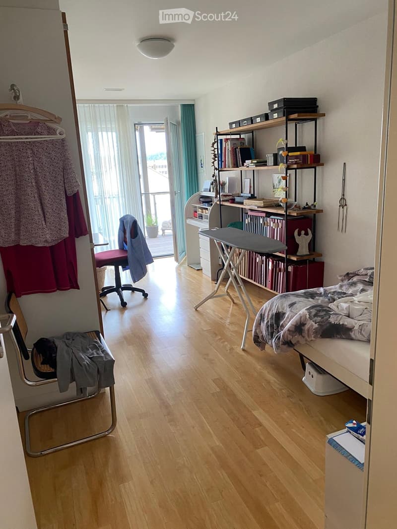 A furnished bedroom with a bed, chair, shelves, ironing board, and storage boxes. The room features wooden flooring and a balcony access.
