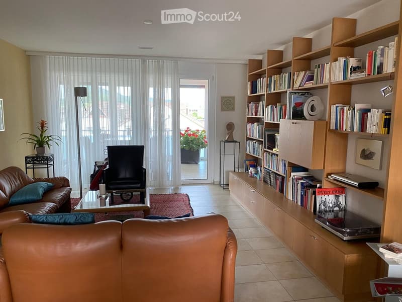 Modern living room with leather sofa, armchair, coffee table, bookshelf filled with books, laptop, and decorative plant. Large windows with sheer curtains and a view of the balcony.