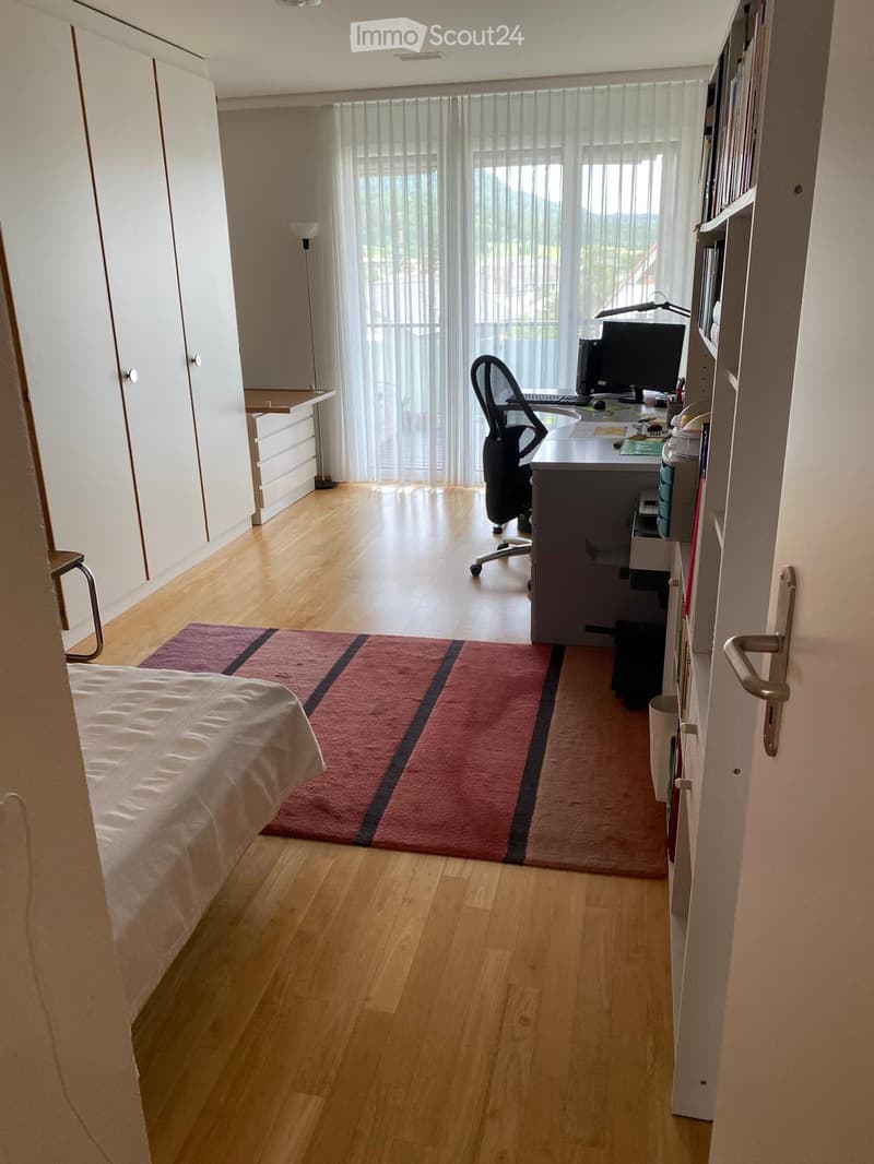 Bright bedroom with a bed, desk, chair, bookshelf, and a colorful rug. Large windows with sheer curtains let in natural light.