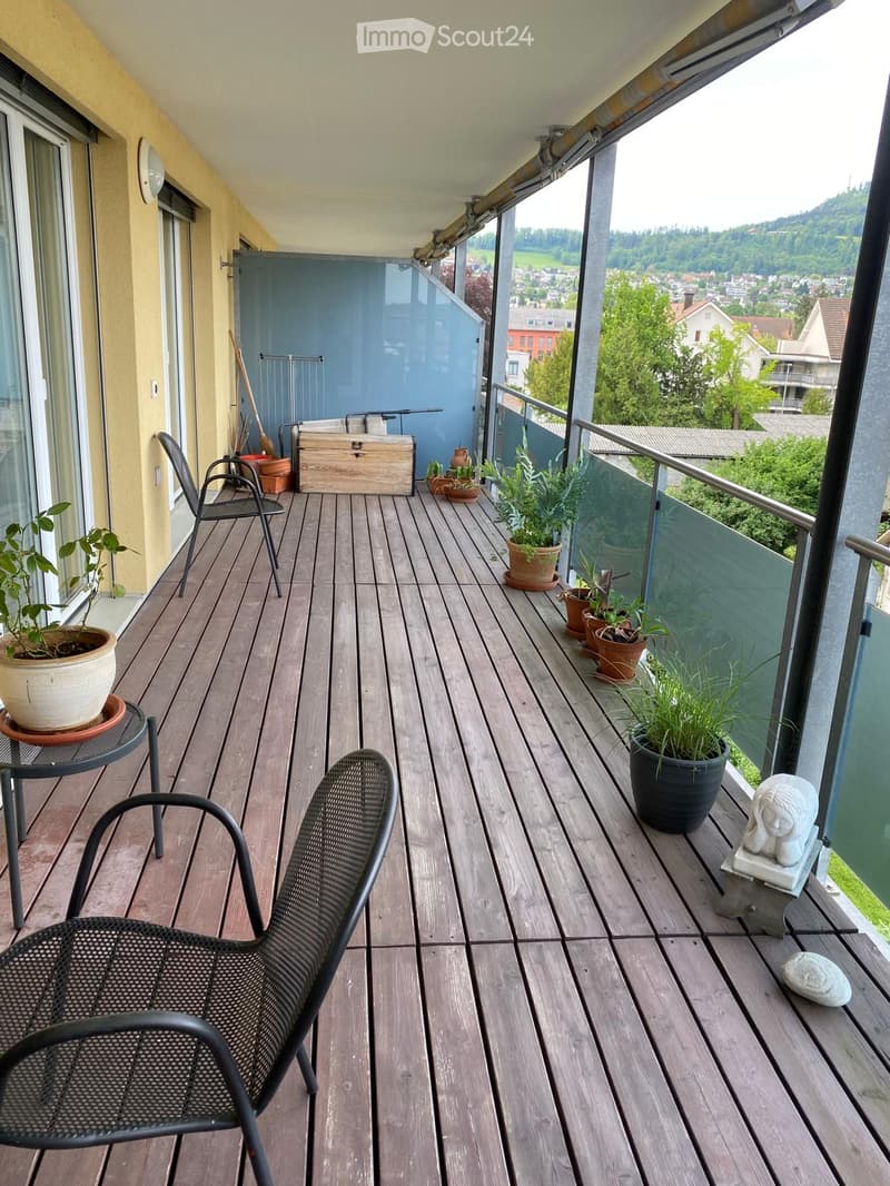 Wooden balcony with a view, featuring a black metal chair, a small table, several potted plants, and a decorative statue.