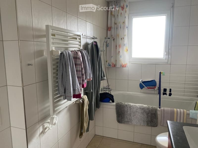 Bathroom with a bathtub, toilet, towel rack, and various towels. The room features tiled walls and a window with a curtain.