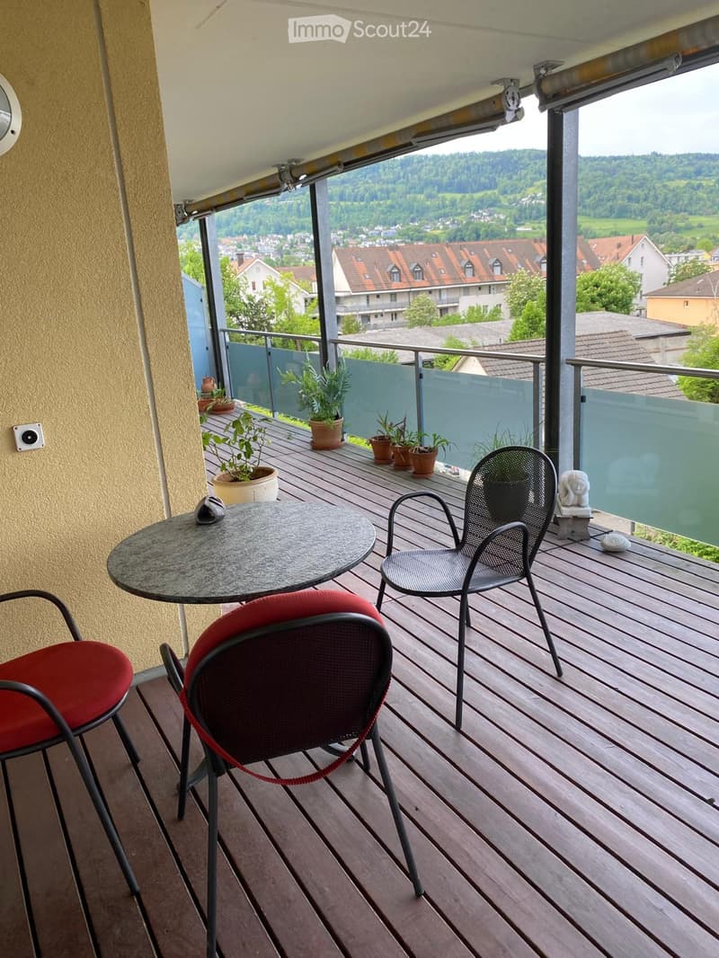 Balcony with wooden decking, round table, three chairs, several potted plants, and a scenic view of the town and surrounding landscape.