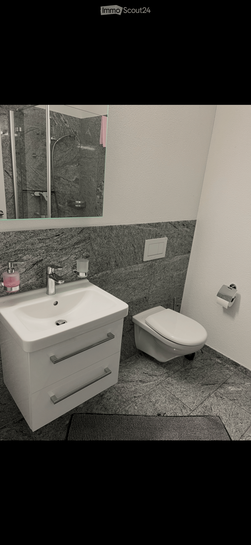 Modern bathroom with a white sink, chrome faucet, toilet, and a large mirror above the sink. The walls are tiled in a dark grey marble pattern, and the floor is covered with matching tiles.