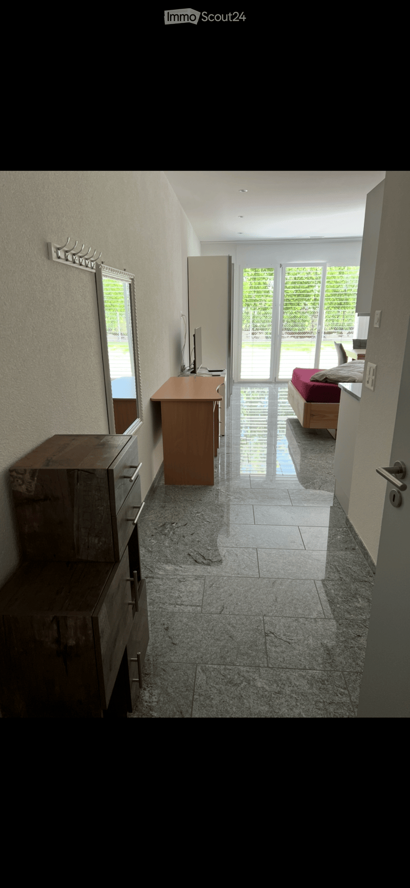 Hallway leading to a bedroom with a bed, a table, and a mirror. The floor is tiled, and the walls are light-colored.