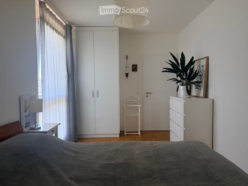 A modern bedroom with a large bed, nightstand, lamp, wardrobe, plant, picture frame, and a small chair near the door.