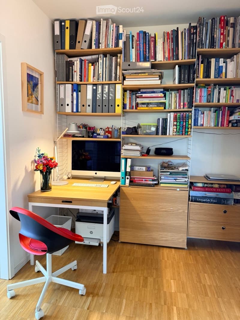 A modern, well-organized home office with a wooden desk, ergonomic chair, large bookshelf filled with books and binders, laptop, printer, and decorative vase with flowers.