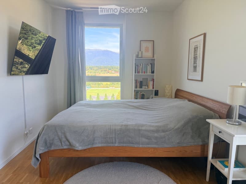 Cozy bedroom with a wooden bed, nightstand, lamp, bookshelf, and large window with a scenic mountain view.