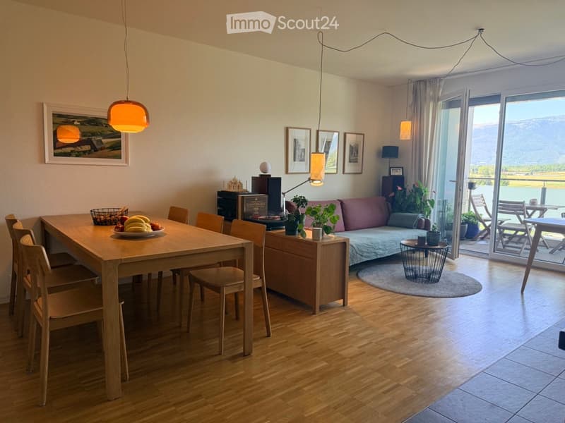 Bright living and dining area with wooden flooring, a sofa, dining table with chairs, pendant lights, and a view of a lake through large windows and sliding doors.