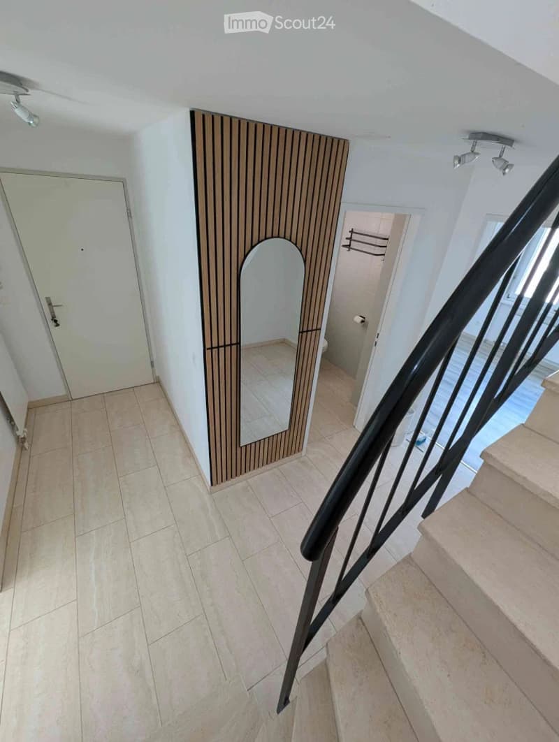 Hallway with tiled floor, modern black staircase railing, and a small adjacent bathroom with a toilet and towel rack.