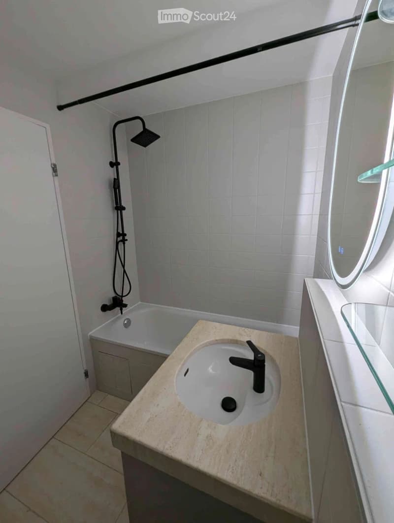 Modern bathroom with a white tiled shower area, a bathtub, a stone countertop sink with a black faucet, and a large round mirror.