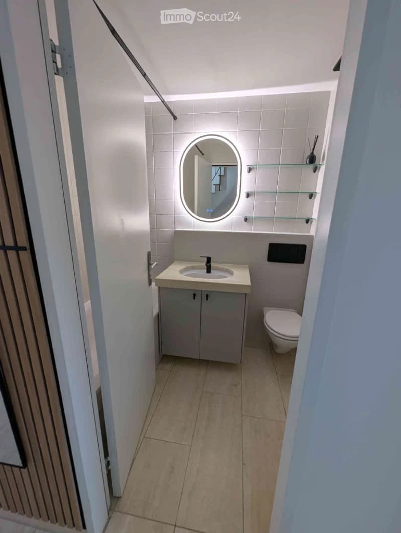 Modern bathroom with a white tiled wall, a round illuminated mirror, a sink with a cabinet below, and a toilet. Glass shelves are mounted on the wall.