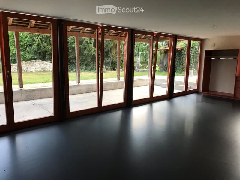 An empty room with dark flooring and large sliding glass doors leading to an outdoor area with a stone wall and greenery.