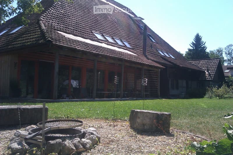 A modern house with a large, well-maintained lawn and a stone fire pit in the foreground. The house features a red-tiled roof, large windows, and a covered patio area with outdoor furniture.