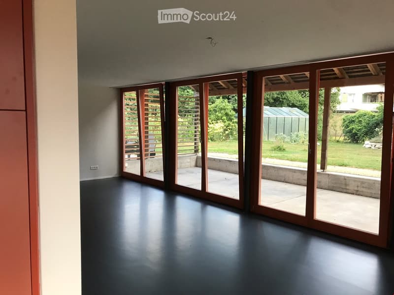Empty room with dark flooring and large sliding glass doors leading to an outdoor patio area with greenery.