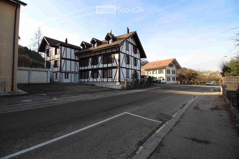 A two-story building with a traditional half-timbered facade and a tiled roof, situated on a quiet street with a parking area in front.
