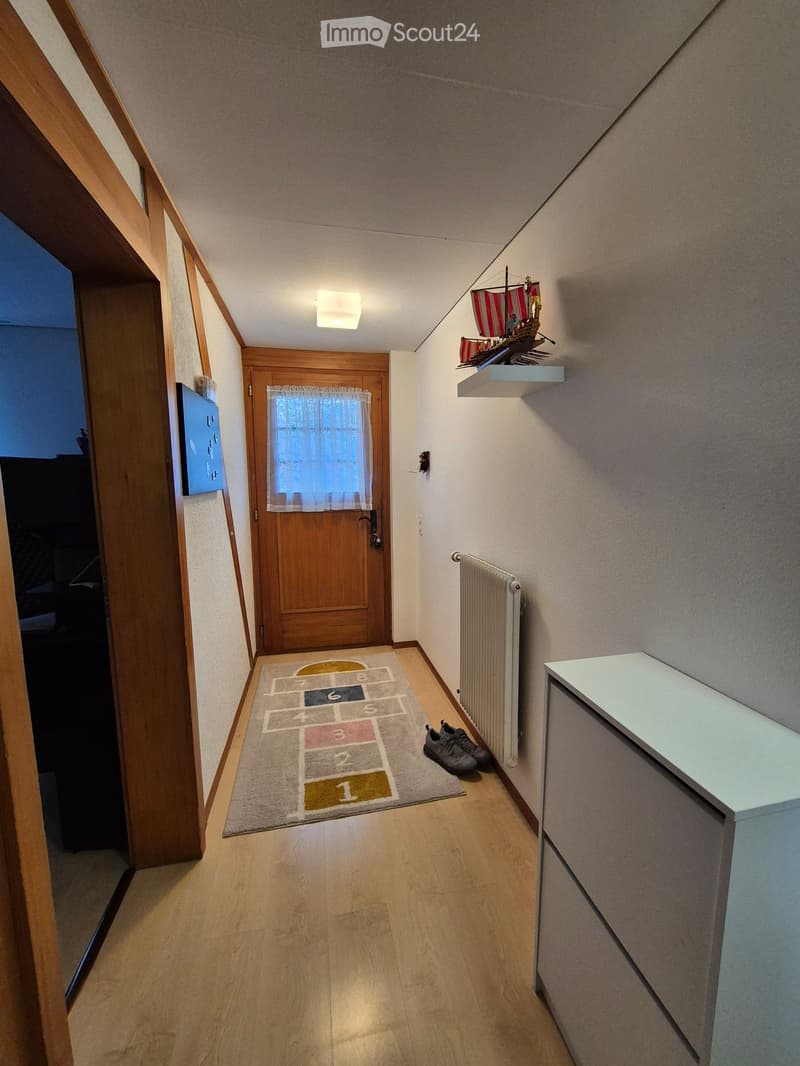 Narrow hallway with wooden flooring, a small rug with numbered squares, a pair of shoes, a radiator, and a shelf with a decorative model ship.