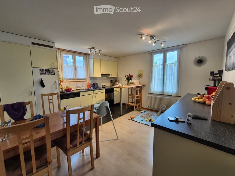 Bright kitchen with light-colored cabinets, a dining area with a wooden table and chairs, and a counter with various items including a vase with flowers, fruits, and a small teddy bear. The kitchen features modern appliances, ample natural light from larg