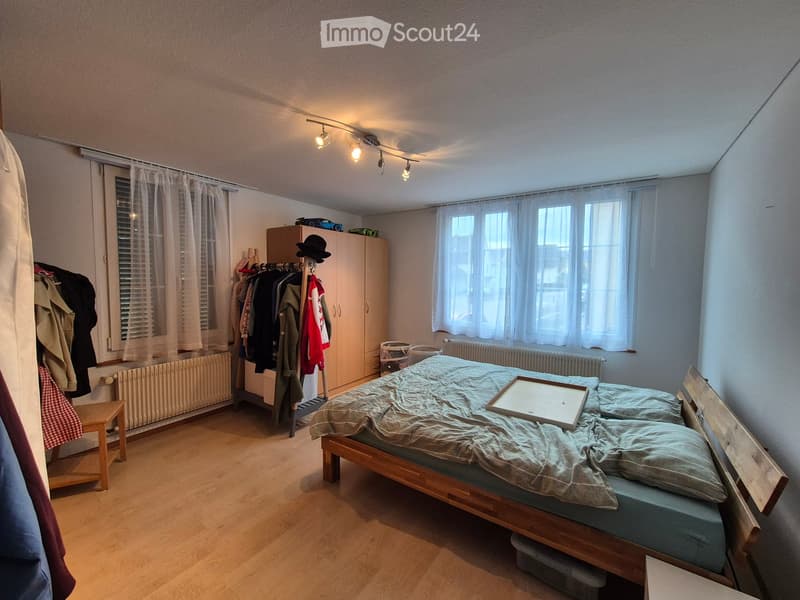Bedroom with wooden floor, bed with pillows and blanket, wardrobe, stool, and curtains on windows.