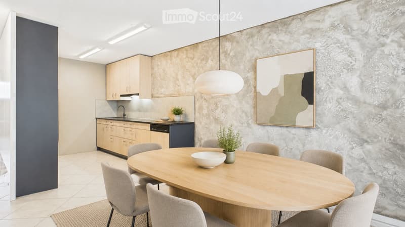 Modern open-plan kitchen and dining area with light wood cabinets, a round wooden dining table surrounded by chairs, a pendant light, and a large mirror on the textured wall.