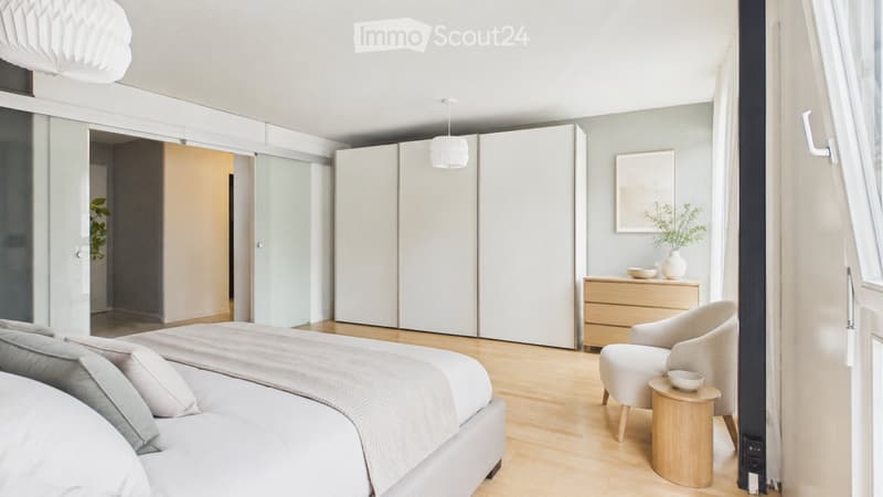 Modern bedroom with a large bed, nightstand, armchair, small table, and a wardrobe. The room features light wood flooring and large windows with sheer curtains.