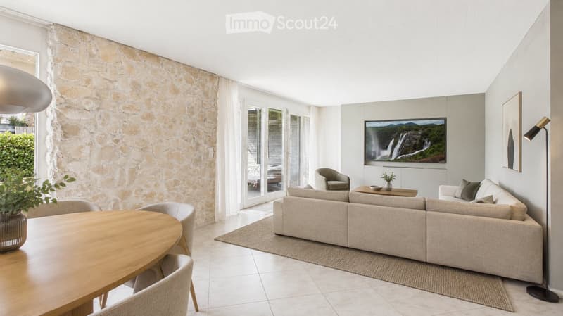 Modern living room with a stone accent wall, large windows, a beige sofa, coffee table, dining table with chairs, and a television displaying a nature scene.