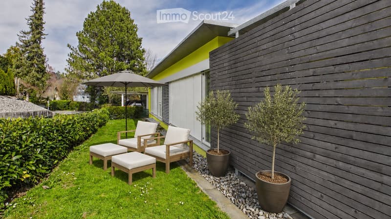 A modern outdoor patio area with white cushioned chairs, matching stools, a large umbrella, and potted trees near a wooden wall.
