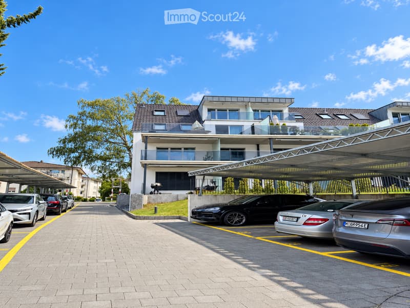 Modern multi-story residential building with balconies, surrounded by a paved parking area with several parked cars under covered structures.