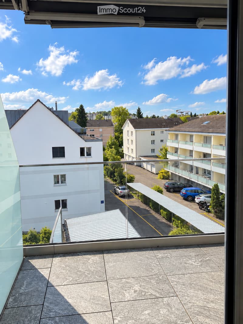 Balcony with tiled floor, metal railing, overlooking a parking area with several cars and surrounding buildings.