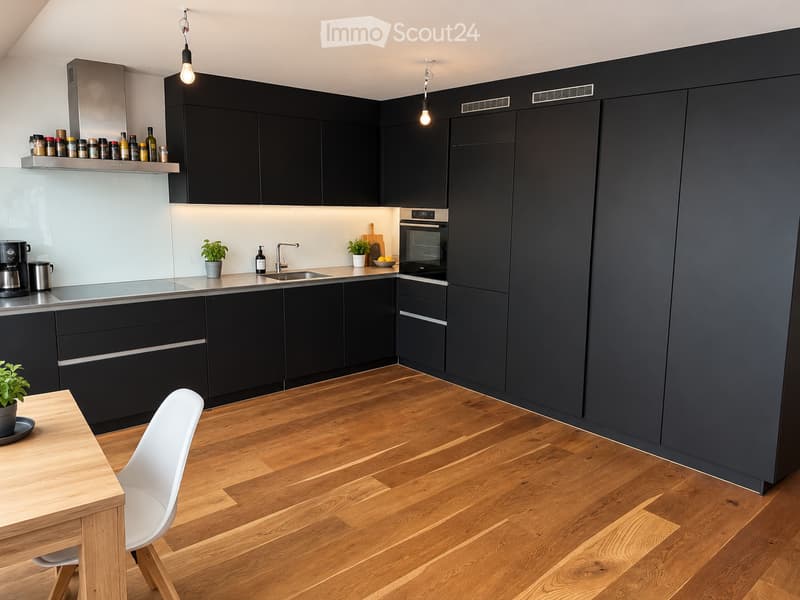 Modern kitchen with dark cabinets, wooden flooring, and stainless steel appliances. Features a dining table with a chair, a coffee maker, and potted plants on the counter.