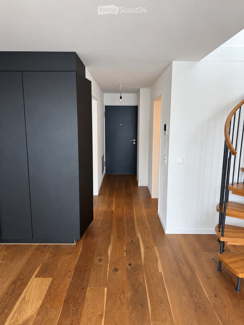 Modern hallway with light wooden flooring, white walls, and a black cabinet on the left. A wooden staircase with a metal railing is visible on the right, leading upwards.