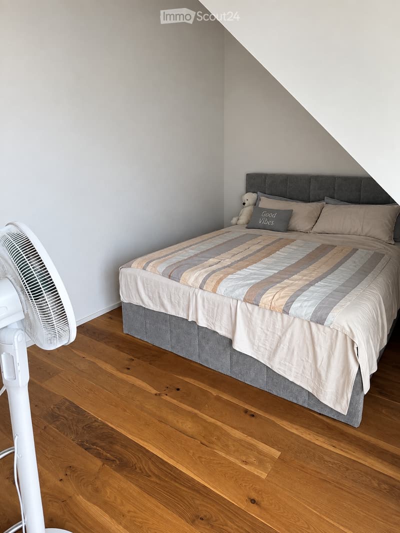A cozy bedroom with a wooden floor, a large bed with a grey headboard, a striped duvet, pillows, and a small teddy bear toy. A white fan is positioned on the left side of the room.