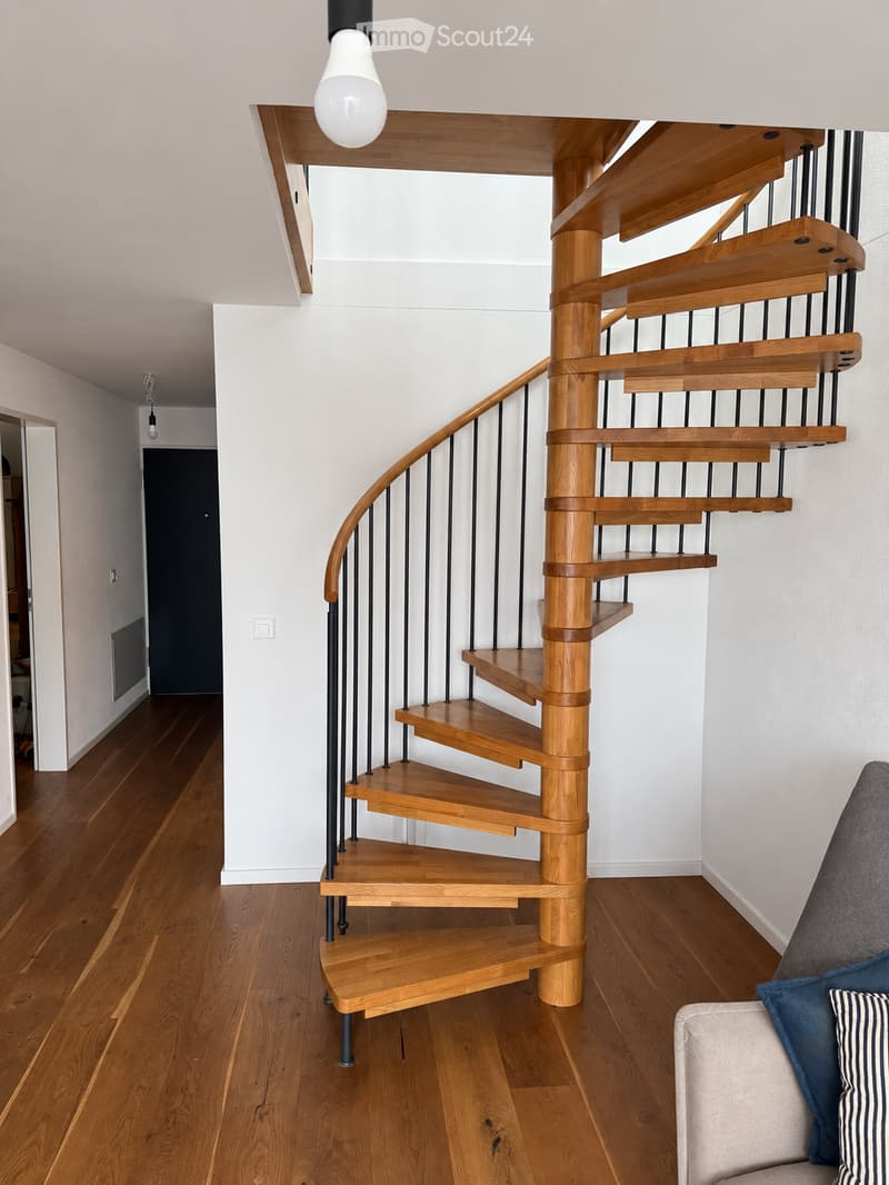 Spiral wooden staircase with black metal railing, wooden flooring, white walls, and a visible light fixture above.