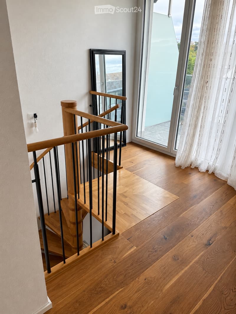 Spiral staircase with wooden steps and a black metal railing, leading up to a landing area with wooden flooring and a large window with white curtains.