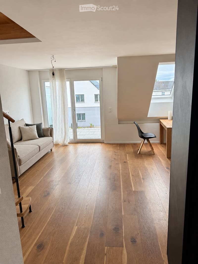 Bright living room with wooden flooring, large windows, and a sliding glass door leading to a balcony. Features a sofa, a modern chair, and a small table.