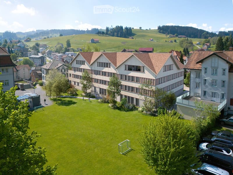 A multi-story building with a red-tiled roof and white walls, surrounded by a well-maintained lawn, trees, and parked cars. The building is part of a larger complex with additional structures visible in the background, set in a rural, hilly landscape.