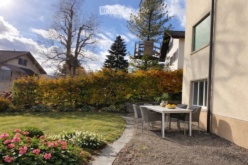 Garden with a patio area featuring a white table and four chairs, surrounded by greenery and flowers, with a pathway leading to the lawn.