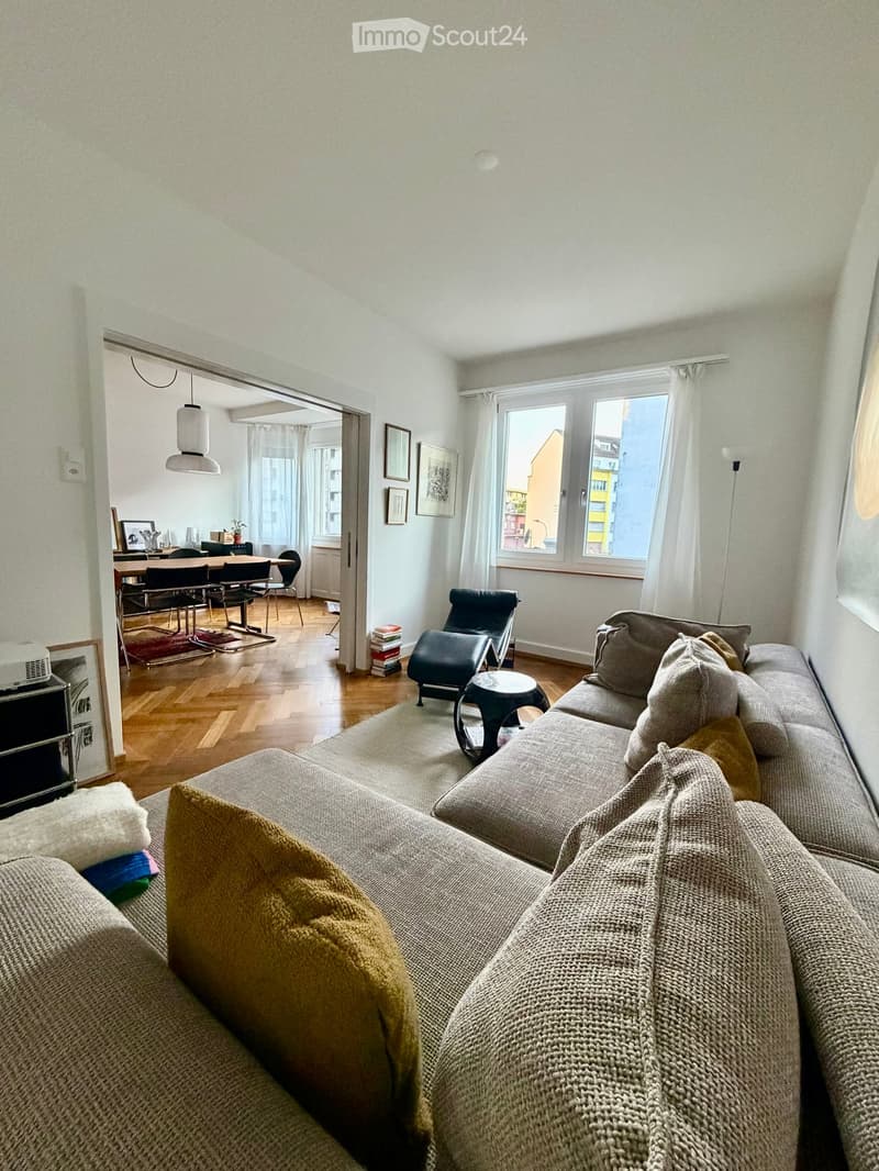 A modern living room with a grey sofa, coffee table, and a dining area with a table and chairs. The room features large windows with white curtains and a view of the city.