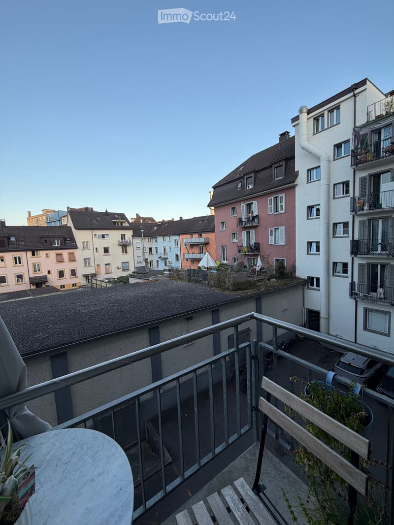 Balcony with a metal railing, a round table, and a wooden chair, overlooking a cityscape with colorful buildings and rooftops.