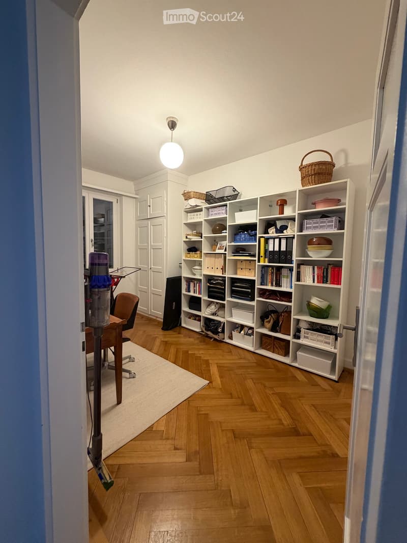 A well-organized room with wooden herringbone flooring, a white bookshelf filled with books and storage boxes, a small table with a chair, and a hanging lamp.