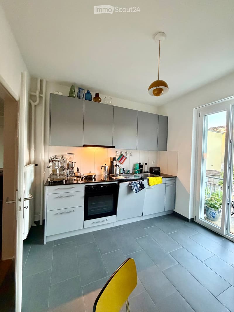 Modern kitchen with grey cabinets, stainless steel appliances, and a black countertop. Features a coffee machine, blender, and various kitchen utensils. A yellow chair is visible in the foreground, and a sliding glass door leads to a balcony with plants.