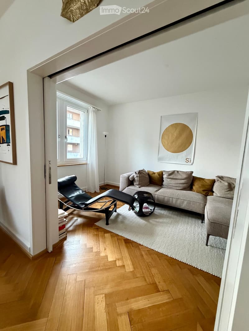 Modern living room with light wooden flooring, a grey sofa with multiple cushions, a black coffee table, a black leather chair, a floor lamp, and artwork on the walls.