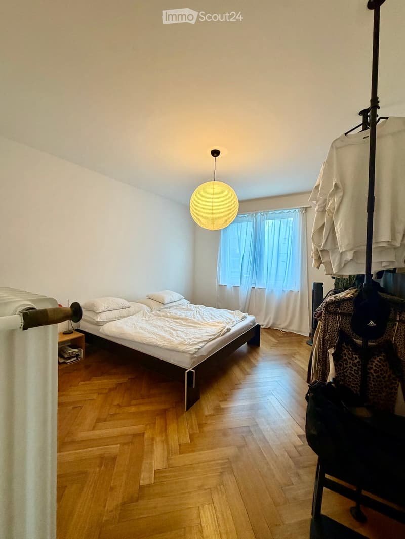 Cozy bedroom with wooden flooring, a double bed with white bedding, a nightstand with a lamp, and a clothing rack with folded shirts and hanging items.