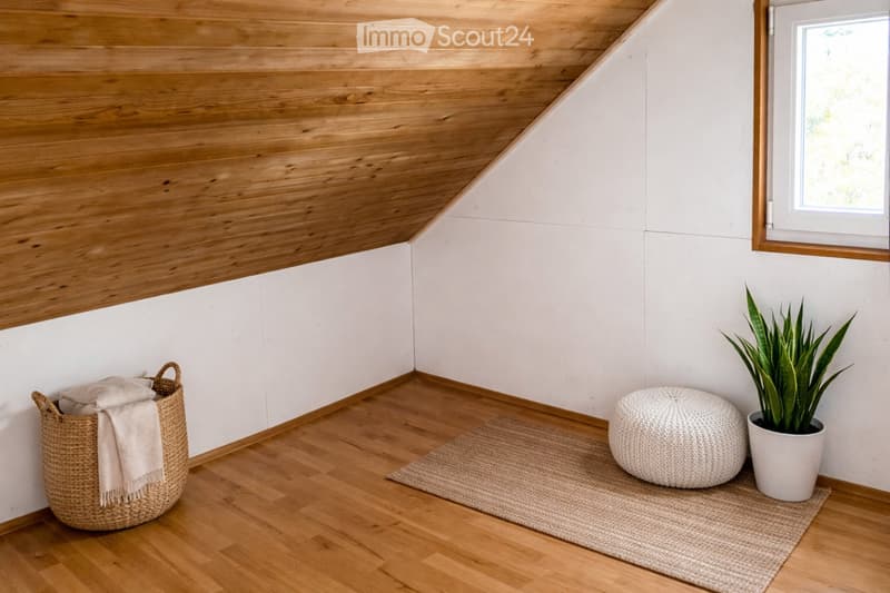 A cozy, unfurnished room with wooden floors, white walls, and a slanted wooden ceiling. Features include a wicker basket with a blanket, a round ottoman, a small rug, and a potted plant near a window.